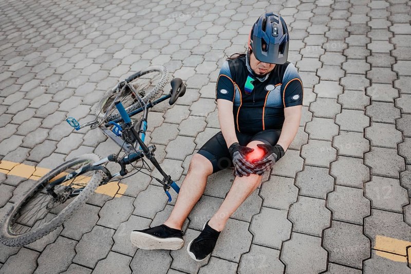 Unhappy cyclist sitting on the pavement with knee pain. Concept of a cyclist with knee injury outdoors, Cyclist with knee pain outdoors
