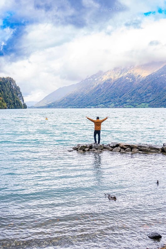 Person standing with outstretched arms on rocks by the lake with mountain backdrop under dramatic sky, Lake Brienz, Switzerland, Europe