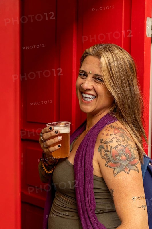 Smiling tattooed woman holding a plastic cup of beer, wearing a purple shawl and a bindi, standing at the entrance of a red door.