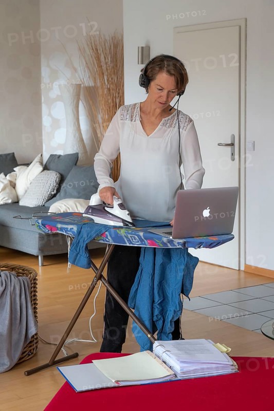 Woman, mid-50s, works from home, with laptop and communicates with colleagues via headset, home office, irons laundry in parallel