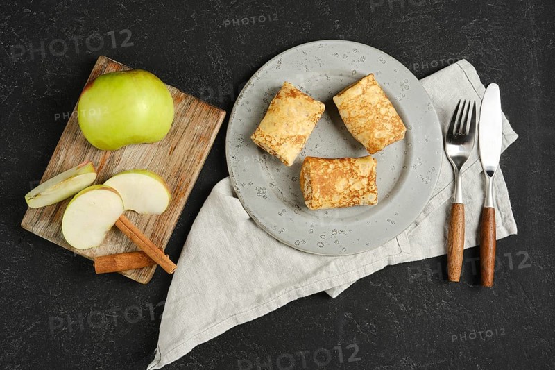 Three golden crepes filled with apples are arranged on a plate. Slices of green apple and a stick of cinnamon accompany the dish, all set on a textured dark background.