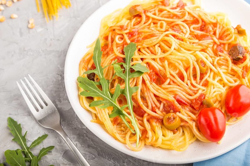 Corn noodles with tomato sauce and arugula on a gray concrete background and blue textile. Side view, close up, selective focus