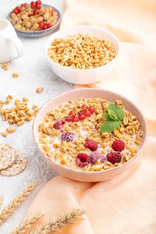 Wheat flakes porridge with milk, raspberry and currant in ceramic bowl on gray concrete background and orange linen textile. Side view, close up