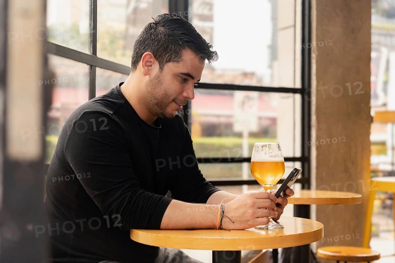 Profile of Latin man sitting enjoying a glass of beer while texting with his smartphone in a pub during the day. The relaxed daytime atmosphere and modern pub environment enhance a casual and enjoyable moment