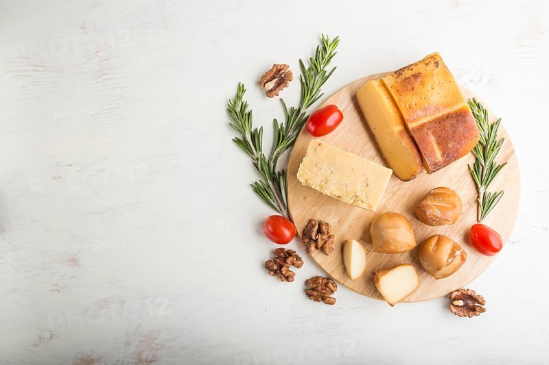 Smoked cheese and various types of cheese with rosemary and tomatoes on wooden board on a white wooden background. Top view, flat lay, copy space