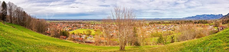 Panoramic view of a village with green meadows and trees, mountains in the background under a cloudy sky, Bad Feilnbach