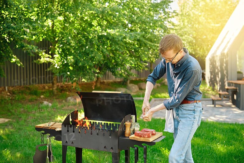 Middle-aged man sprinkles raw steak with sea salt using hand mill while cooking at backyard