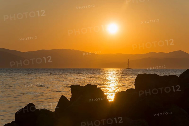 Sunset over the sea with a sailing boat in the distance, rocks in the foreground, Agíos Nikólaos, Agios Nikolaos, Methana peninsula, Argolida regional district, Argolis, Peloponnese, Greece, Europe