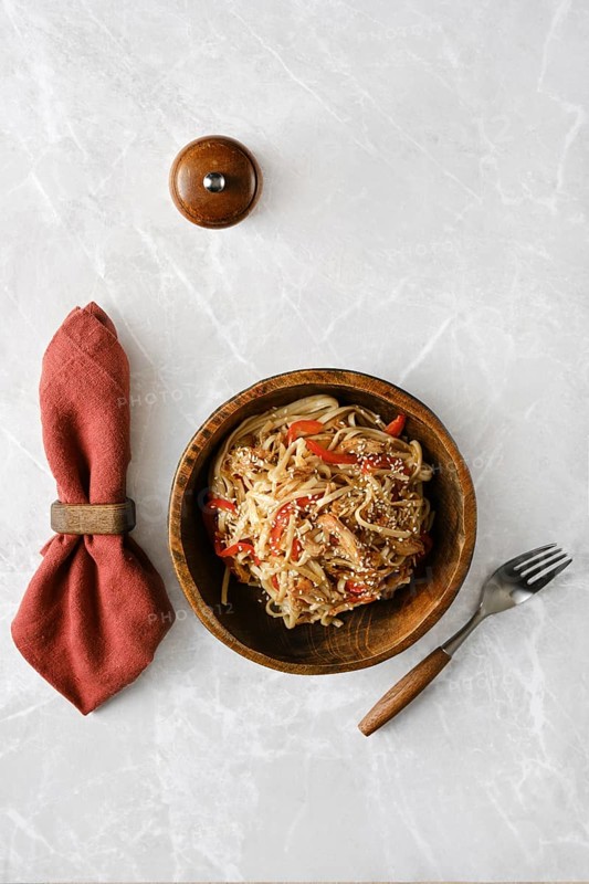 A wooden bowl filled with stir-fried noodles mixed with pulled beef, bell peppers and topped with sesame seeds rests on a marble table, accompanied by a rustic fork and a neatly rolled cloth napkin