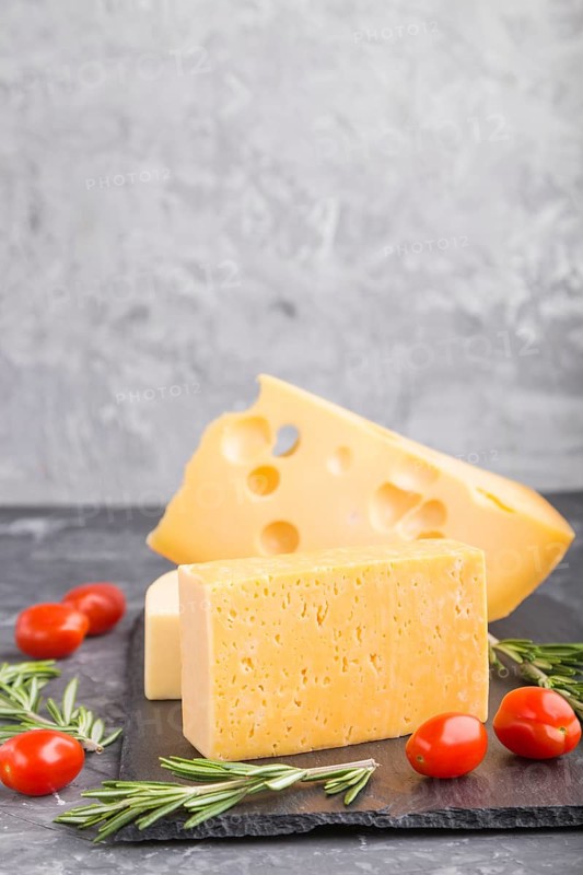 Various types of cheese with rosemary and tomatoes on black slate board on a black concrete background. Side view, close up, copy space, selective focus