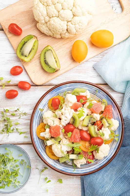 Vegetarian salad of cauliflower cabbage, kiwi, tomatoes, microgreen sprouts on white wooden background and blue linen textile. Top view, flat lay, close up