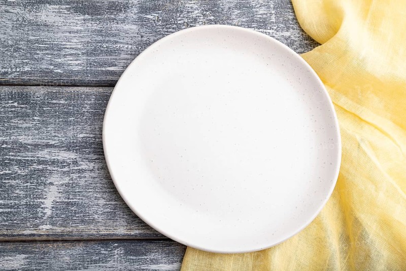 Empty white ceramic plate on gray wooden background and yellow linen textile. Top view, close up, flat lay
