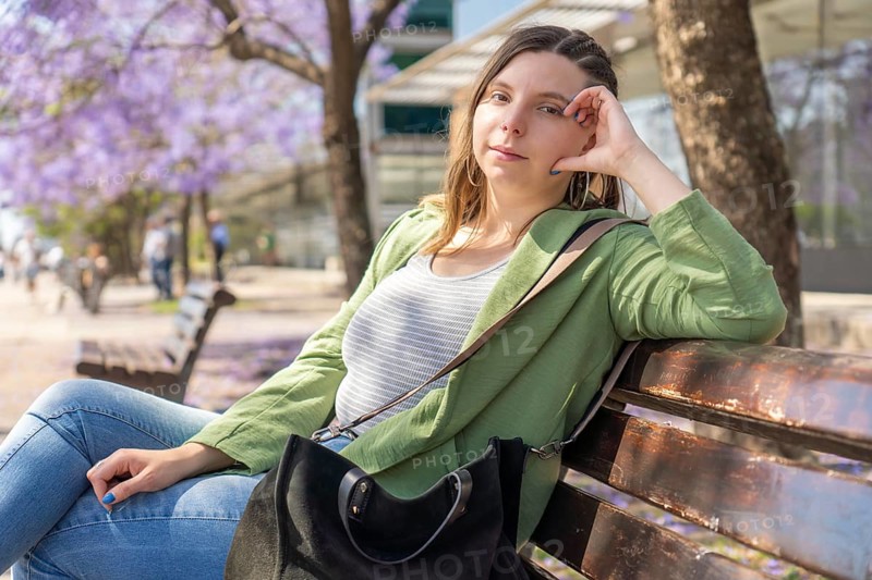 A young woman in a green jacket looks at the camera while sitting relaxed on a bench under purple blooming trees on a sunny day. Blurred people walk in the background, enjoying the pleasant weather
