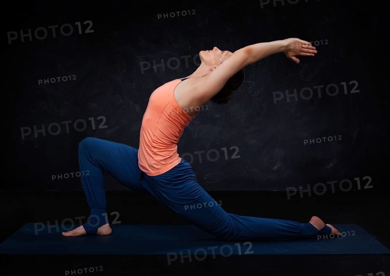 Beautiful sporty fit yogini woman practices yoga asana Anjaneyasana, low crescent lunge pose in surya namaskar on dark background