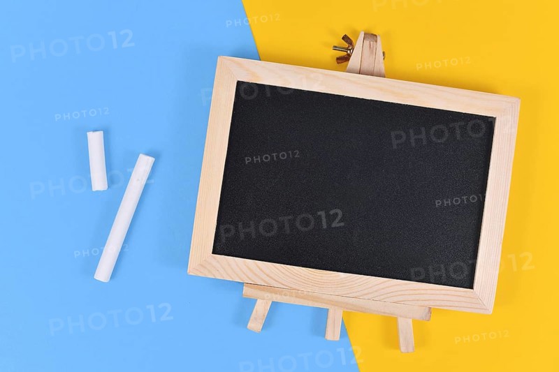 Empty wooden black chalkboard next to pieces of white chalk on yellow and blue background