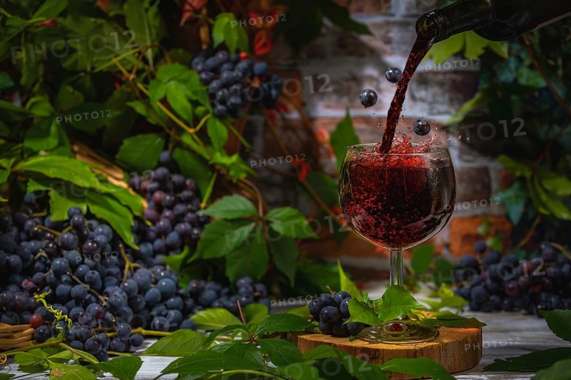 Pouring wine into glass surrounded by blue burgundy grapes in front of brick wall with leaves