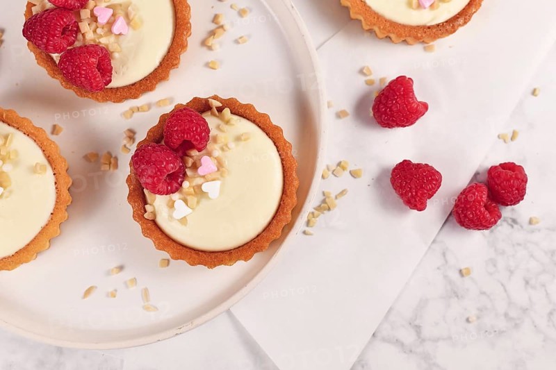 Top view of small tartlet pastries with white cream, topped with raspberry fruits and almond sprinkles