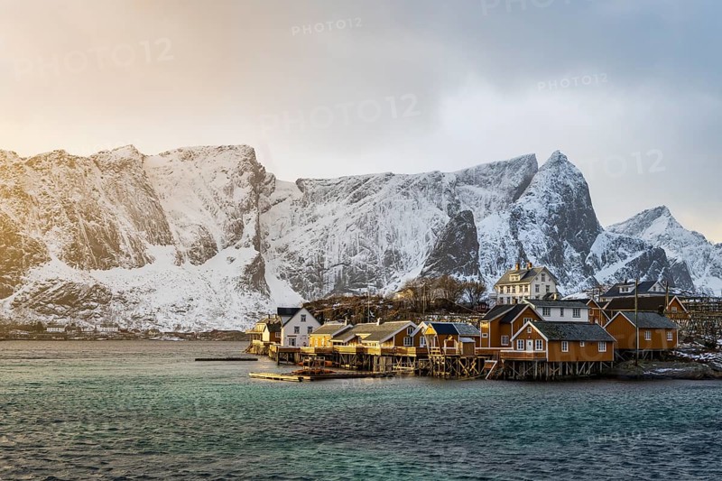 Traditional yellow rorbuer cabins on Sakrisøy Island, snow-capped rocky mountains in the back, Sakrisoy, Reine, Lofoten, Norway, Europe