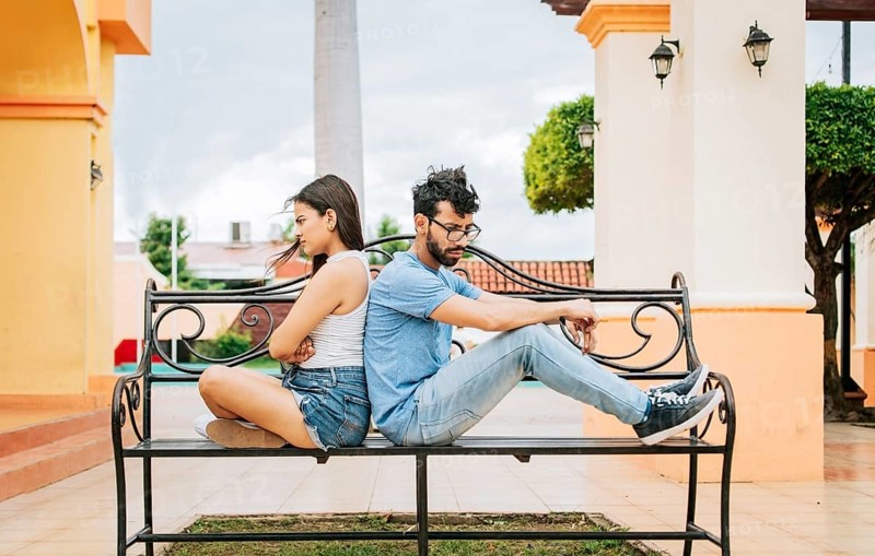 Upset couple sitting back to back on a park bench. Concept of a couple arguing in a park. Upset young couple sitting back to back outside, young couple arguing sitting back to back