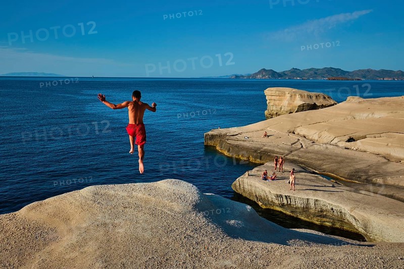 Sarakiniko, Greece, May 23, 2019: Young tourists jump off the white rocks cliff on famous tourist attraction of Milos island Sarakiniko beach and dive into Aegean sea, Milos island, Greece, Europe