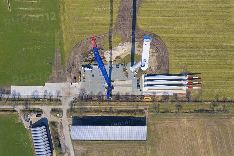 Aerial view over construction site showing rotor blades on the ground and building crane assembling steel tower sections of wind turbine at windfarm