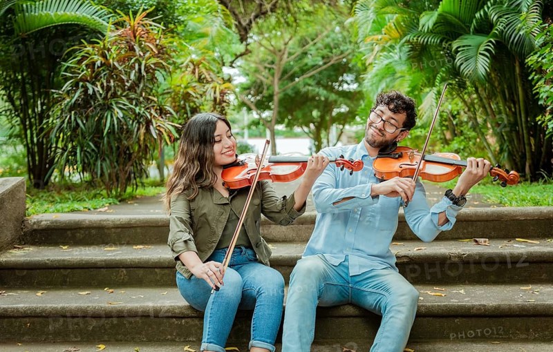 Portrait of two young violists playing violin sitting on stairs outside. Portrait of smiling violinists sitting on stairs playing violin. Man and woman sitting on stairs playing violin