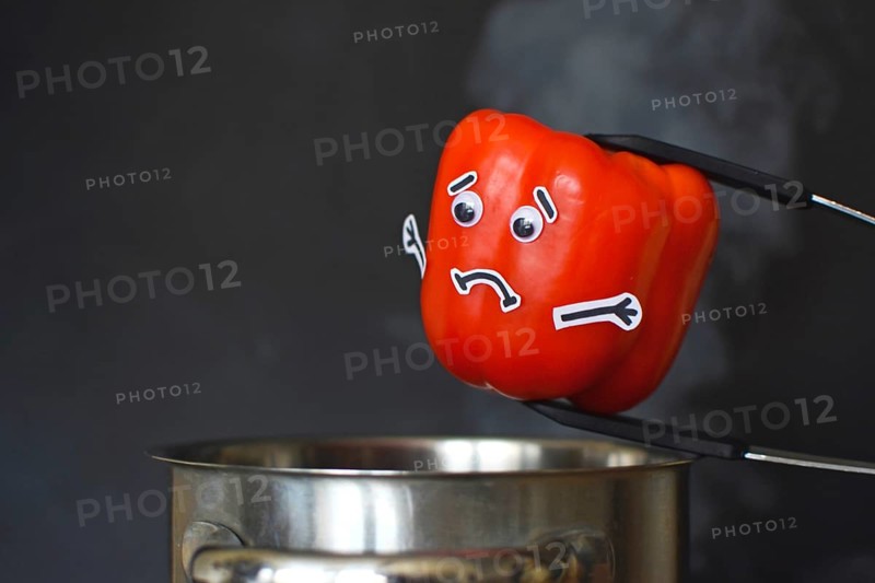 Red Paprika bell pepper with sad face and goggle eyes being put into a steaming cooking pot on dark black background, concept for vegetables losing vitamins when cooked