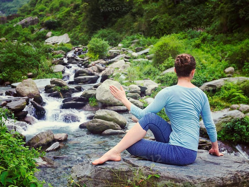 Yoga exercise outdoors, woman doing Ardha matsyendrasana asana, half spinal twist pose at tropical waterfall in Himalayas in India. Vintage retro effect filtered hipster style image
