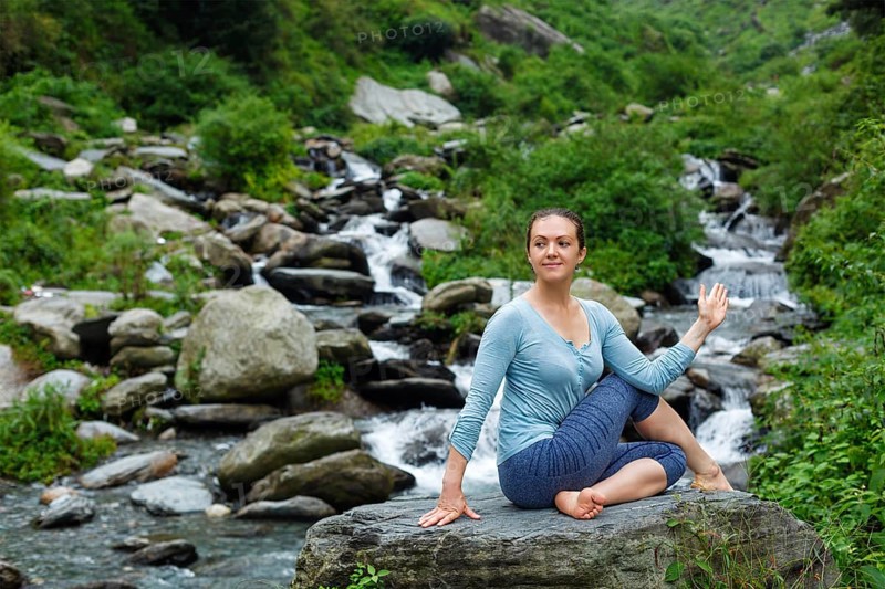 Yoga exercise outdoors, woman doing Ardha matsyendrasana asana, half spinal twist pose at tropical waterfall in Himalayas in India