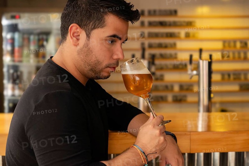 Close-up of a young Latino man savoring a beer in a nightclub. His serious expression and concentrated gaze emphasize his appreciation for the taste, capturing a moment of relaxation and enjoyment amidst the lively nightlife