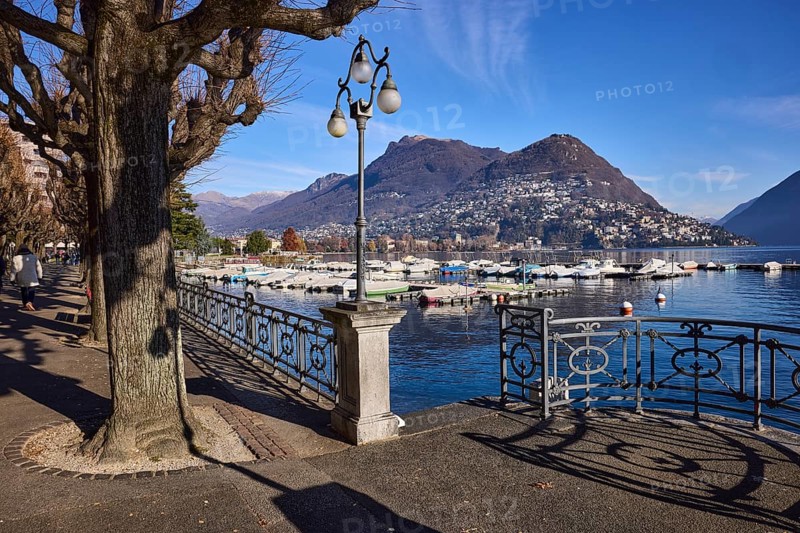 Lantern, railing, trees, promenade, shadow, lake, mountains, blue sky with veil clouds, Lake Lugano, Lugano, district Lugano, canton Ticino, Switzerland, Europe