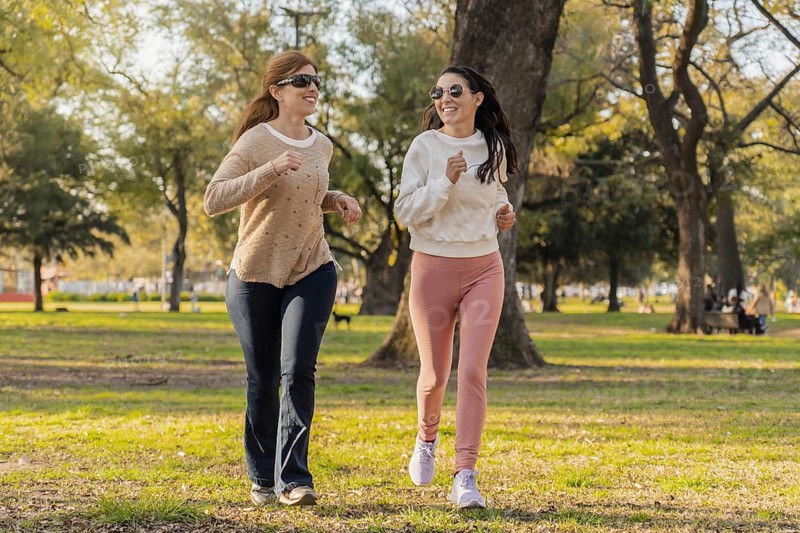 Two women jog together in a sunlit park, enjoying an active lifestyle. They smile as they run through green trees, embracing fitness and health on a beautiful day