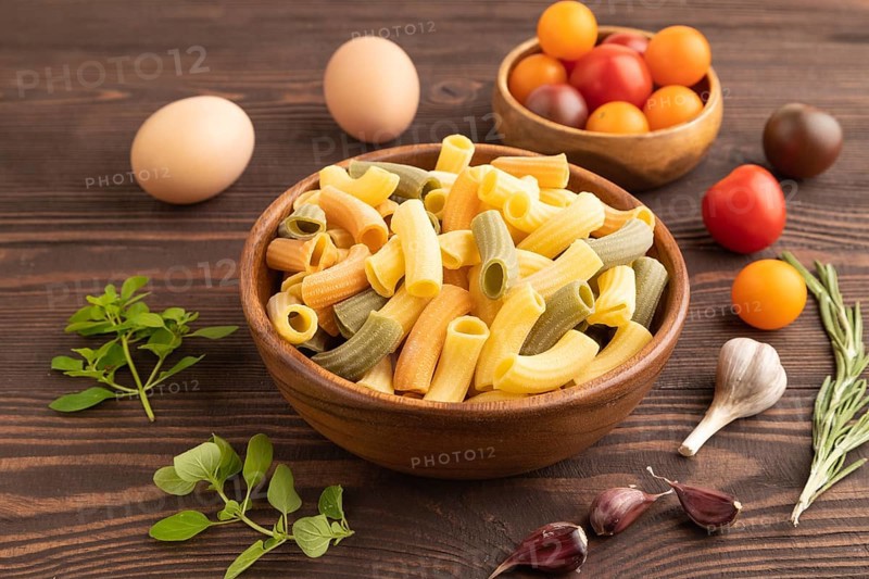 Rigatoni colored raw pasta with tomato, eggs, spices, herbs on brown wooden background. Side view, close up