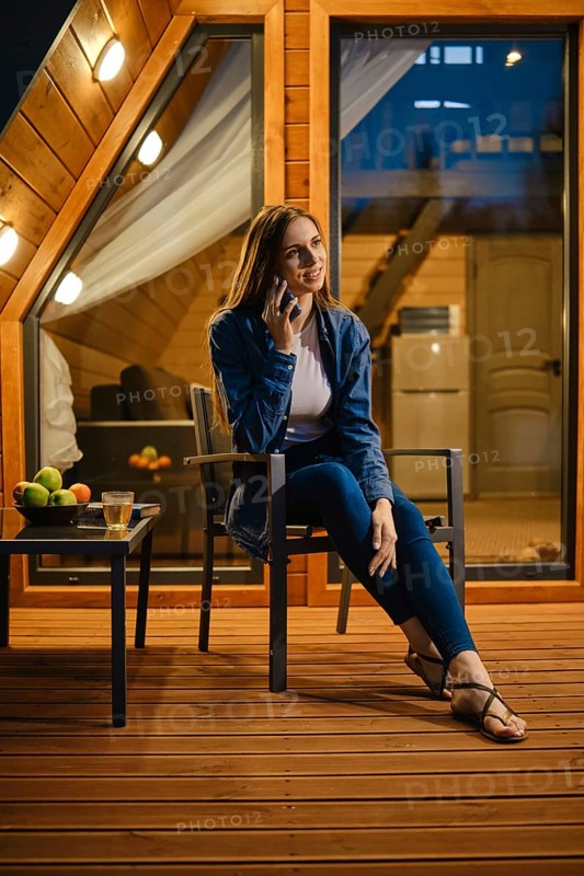 Cute young woman sitting on the terrace of a bungalow late evening and talking on the smartphone
