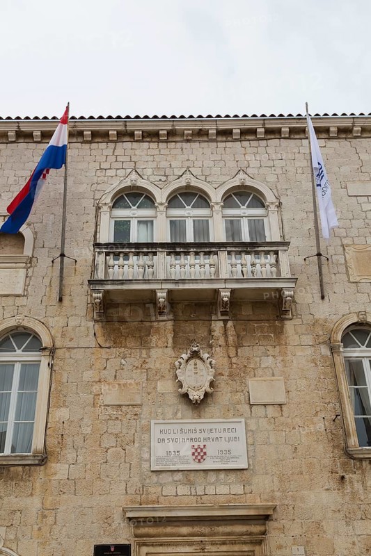 Old beige and tan stone cladded Trogir Town Hall building facade with balcony, flags, arched windows and commemorative 1835-1935 plaque, Trogir, Croatia, Europe