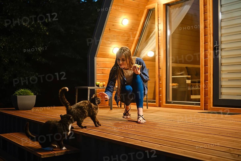 Cute woman sits on a wooden deck and playing with two curious cats