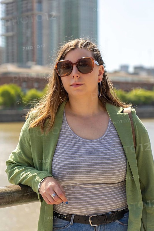 Portrait of a young woman wearing large sunglasses, a striped tank top, and a green jacket, standing near a river with tall buildings in the background on a sunny day