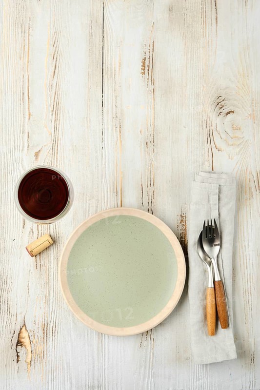 A minimalistic dining arrangement showcases a pale green plate next to a cup of dark beverage, complemented by a fork and knife on a linen napkin against a rustic wooden background