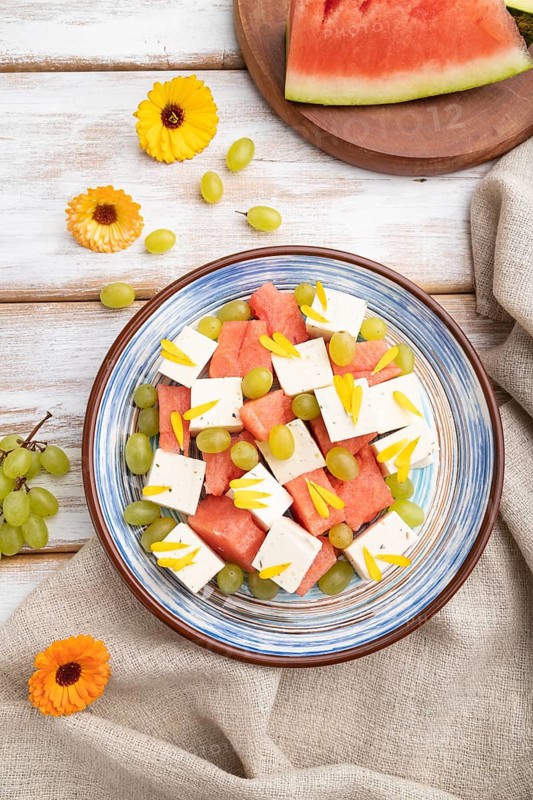 Vegetarian salad with watermelon, feta cheese, and grapes on blue ceramic plate on white wooden background and linen textile. top view, close up, flat lay