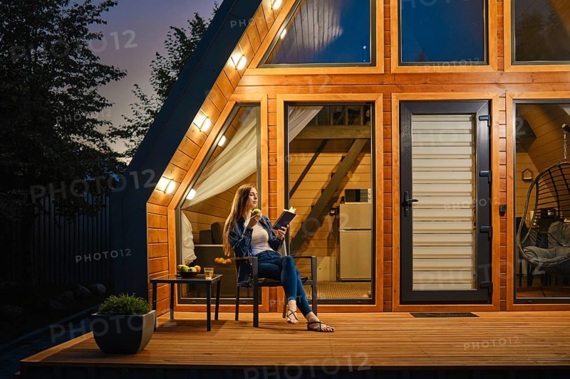 In the silence of the night, a woman sits on the wooden terrace of a modern log cabin and enjoys reading and fresh fruit
