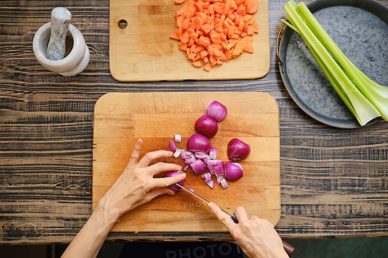 Hands chop red onions on a wooden cutting board with diced carrots and celery nearby. A mortar and pestle sit beside the fresh ingredients in a warm kitchen ambiance