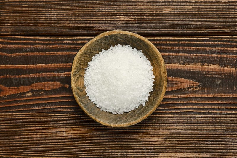 Coarse salt is displayed in a rustic wooden bowl, set against a textured wooden table. The natural materials create a warm, inviting atmosphere, emphasizing culinary simplicity.