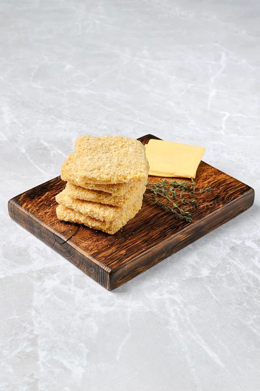 Homemade cheese crackers are neatly stacked on a rustic wooden board. Fresh thyme sprigs accompany the golden crackers and a slice of yellow cheese. This arrangement highlights simplicity and flavor