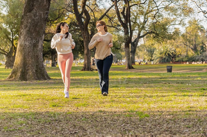 Title:Two women jogging together in a park, embracing fitness and enjoying nature on a sunny dayDescription:Two women are jogging side by side in a sunlit park, enjoying an active lifestyle and each other's company. Surrounded by trees, they smile as they exercise outdoors, promoting health and well-being