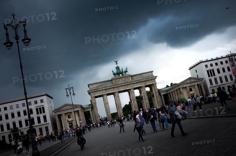 People visit the Brandenburg Gate on a day with a threatening, dramatic sky and heavy clouds, a crow (Corvus corone) flies into the picture, light shines in the background from a gap in the clouds, symbol of Germany, threat, political and economic situation, fear, confidence, hope, early classicism, Triumphtor, archway with quadriga, Pariser Platz, seen from the street Unter den Linden, Berlin Mitte, city of Berlin, Germany, Europe