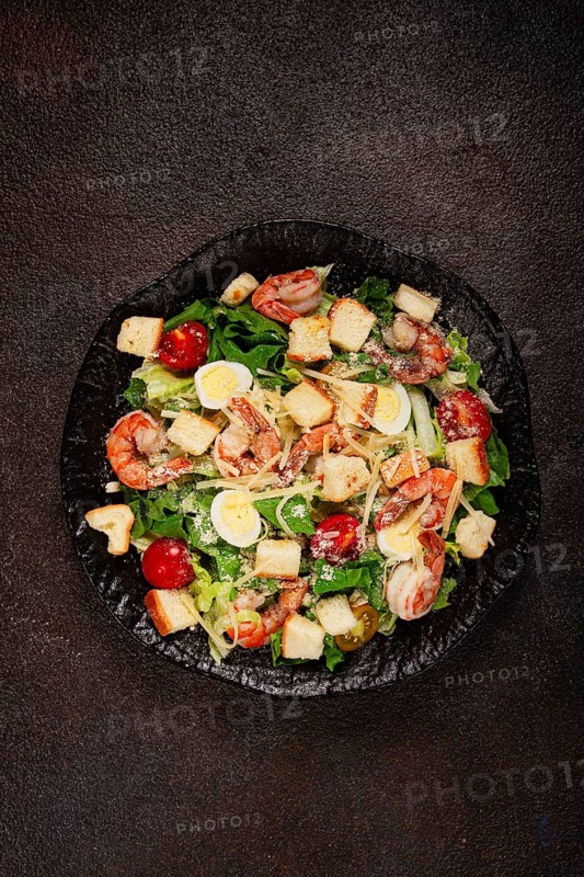 Caesar salad with tiger prawns, shrimp salad, lettuce leaves, quail eggs, cherry tomatoes, parmesan cheese, on a black plate