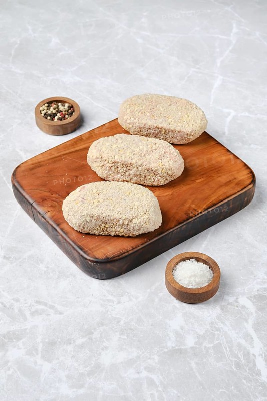 Three breaded cutlets on a rustic wooden cutting board, accompanied by small bowls of salt and mixed spices. The setting highlights a simple yet inviting culinary preparation