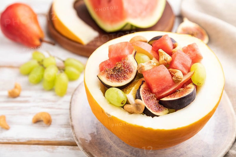 Vegetarian fruit salad of watermelon, grapes, figs, pear, orange, cashew on white wooden background and linen textile. Side view, close up, selective focus