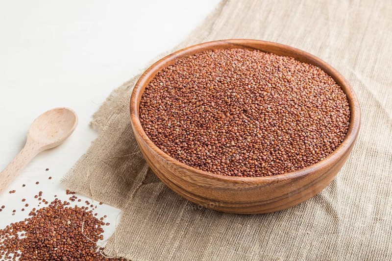 Wooden bowl with raw red quinoa seeds on a white wooden background and linen textile. Side view, close up