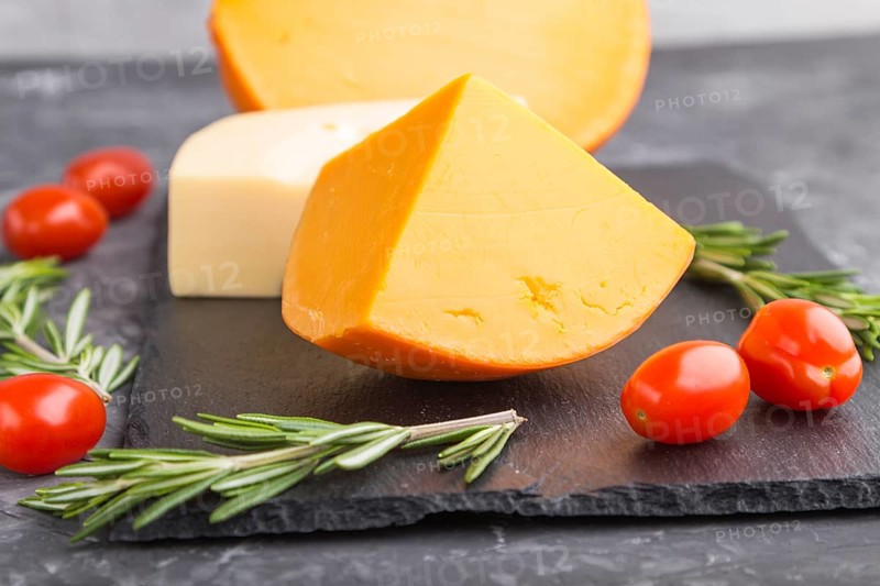 Various types of cheese with rosemary and tomatoes on black slate board on a black concrete background. Side view, close up, selective focus